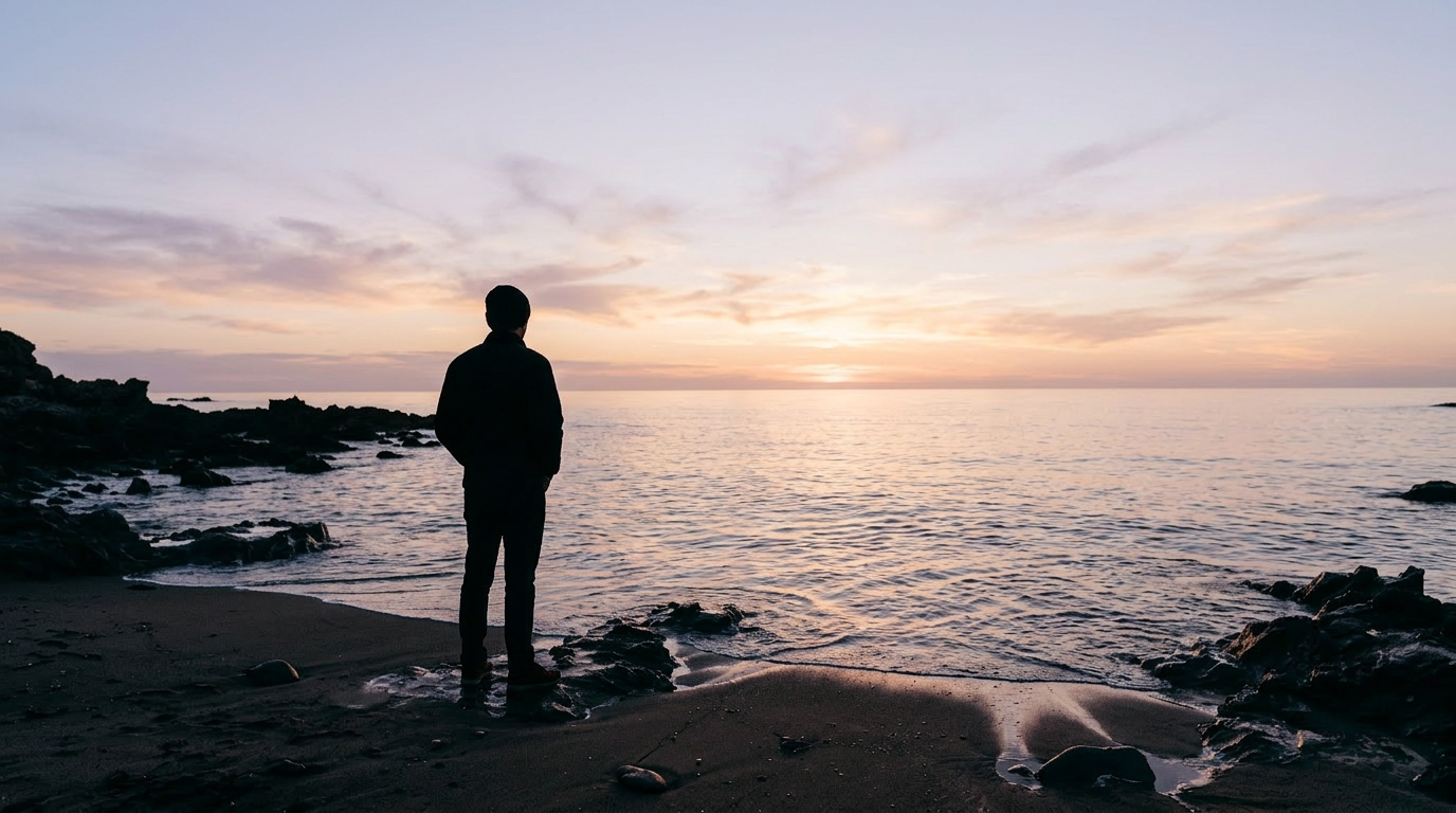 Silhouette of a person standing on a rocky beach at sunset, looking out at the ocean